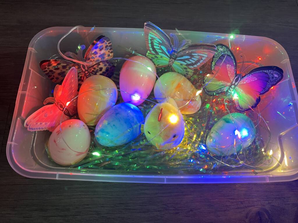 Small container with sensory materials that include 5 plastic butterflies, large plastic eggs with items in it, easter grass, a wind-up chick, and lights.