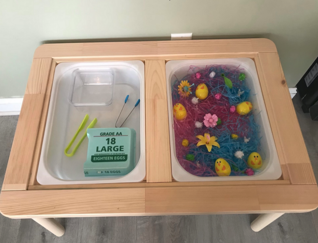 Sensory Bin that is themed towards easter with chick easter eggs, plastic grass, small pompom balls, silk flowers and an empty bin with tongs and an egg container.