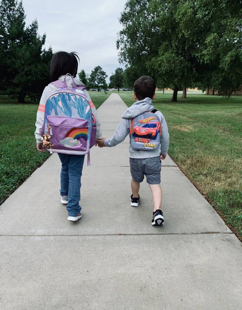Two elementary aged children walking to school holding hands, back packs on. Walking on a sidewalk with a view of them from behind.