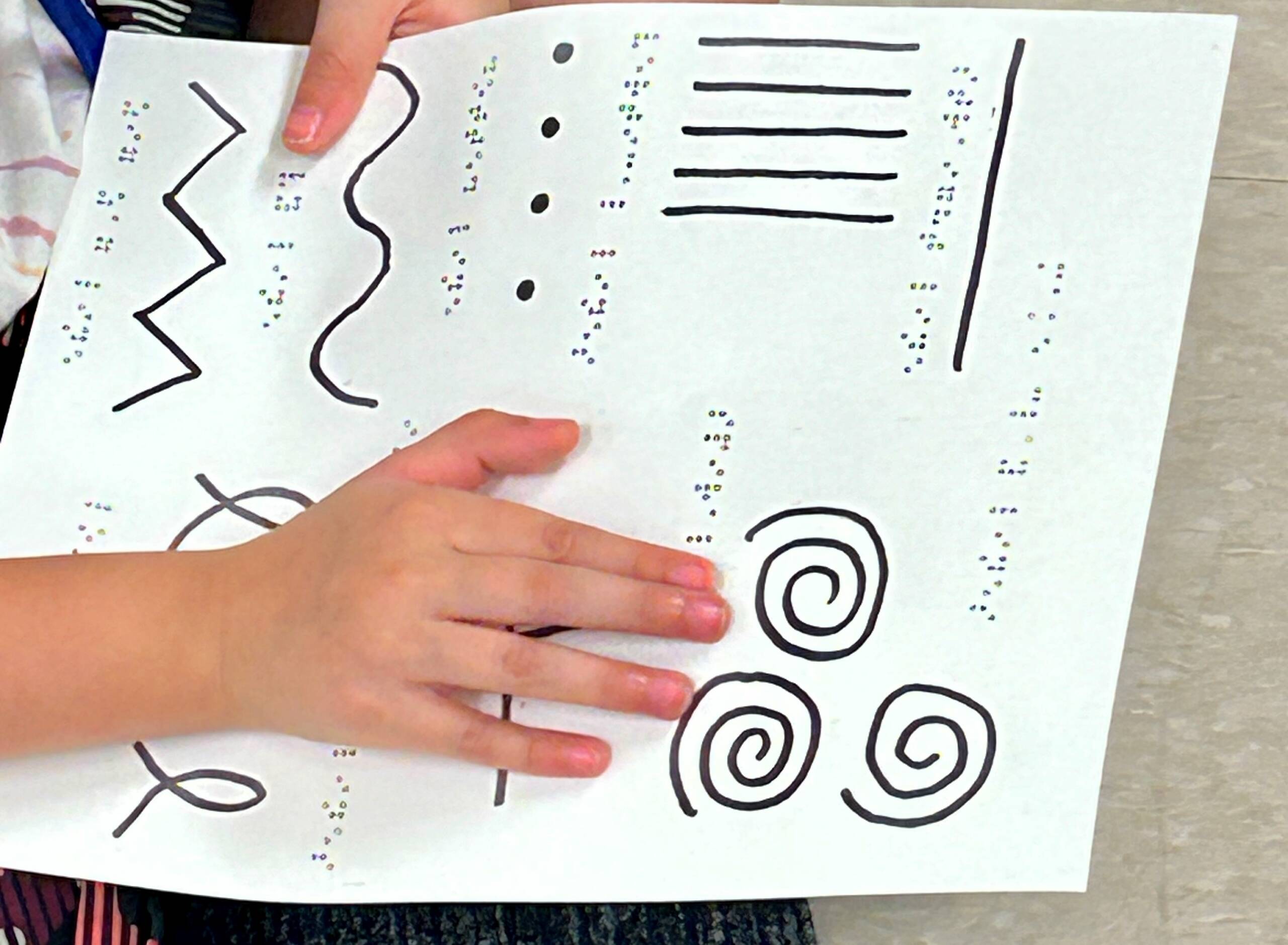 A student holding a piece of paper with a variety of line types such as vertical lines, spirals, a dotted line, and a set of intersecting lines.