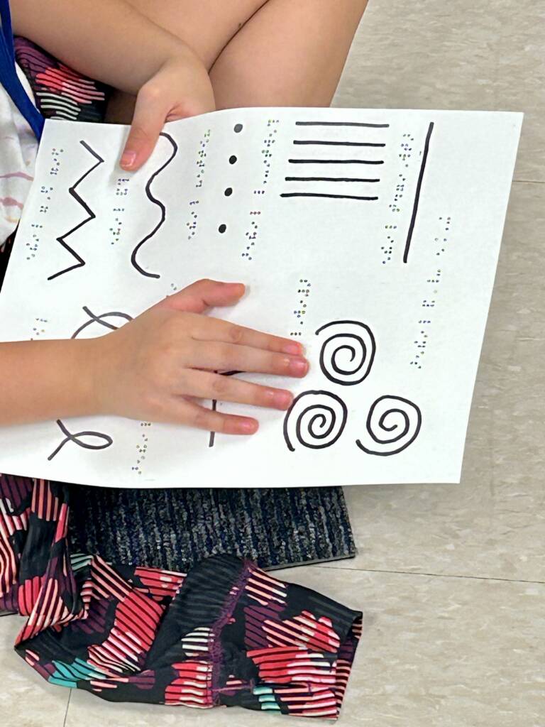 A student holding a piece of paper with a variety of line types such as vertical lines, spirals, a dotted line, and a set of intersecting lines.