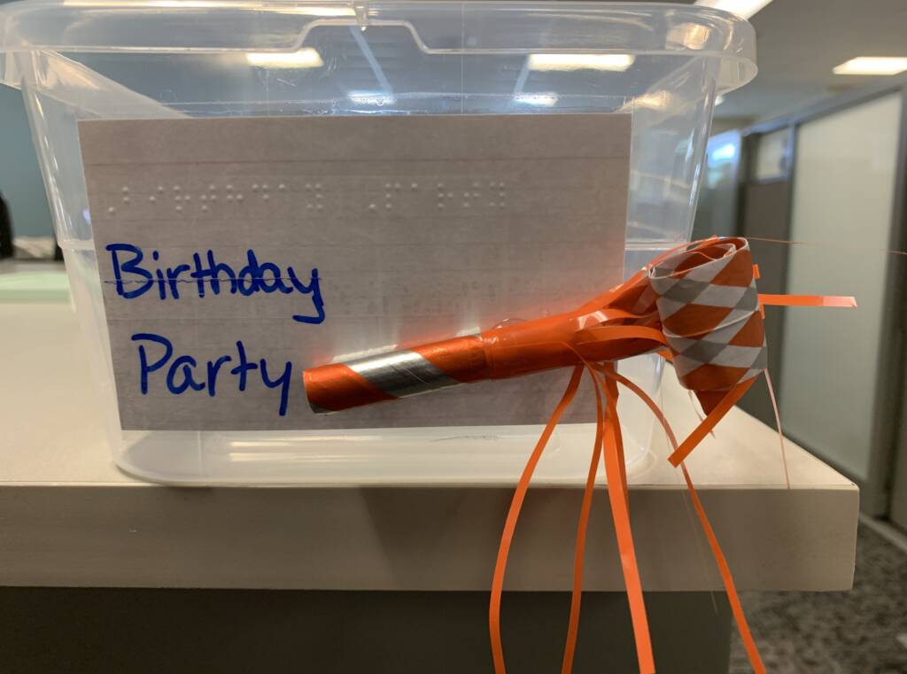 Birthday party container with the with print and braille and a tactile label that includes a paper horn.