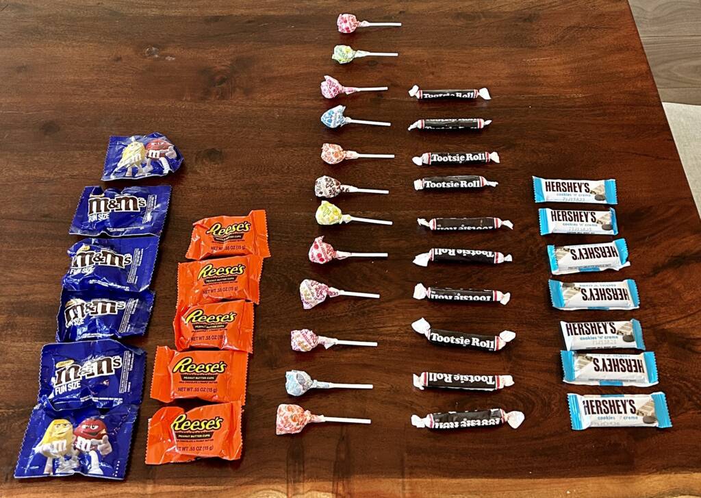 Candy sorted and lined up on the table a row of m&ms, Reese's, lolly pops, tootsie rolls, and snack size candy bars. The lolly pops have the most in their row, followed by; tootsie rolls, m&ms, hershey bars, and last Reese's. 