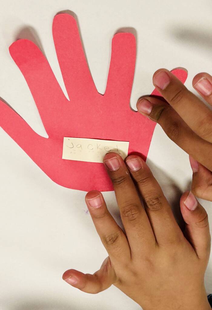 Paper cut out hand with the word jacket in print and braille with the student's hands touching it.