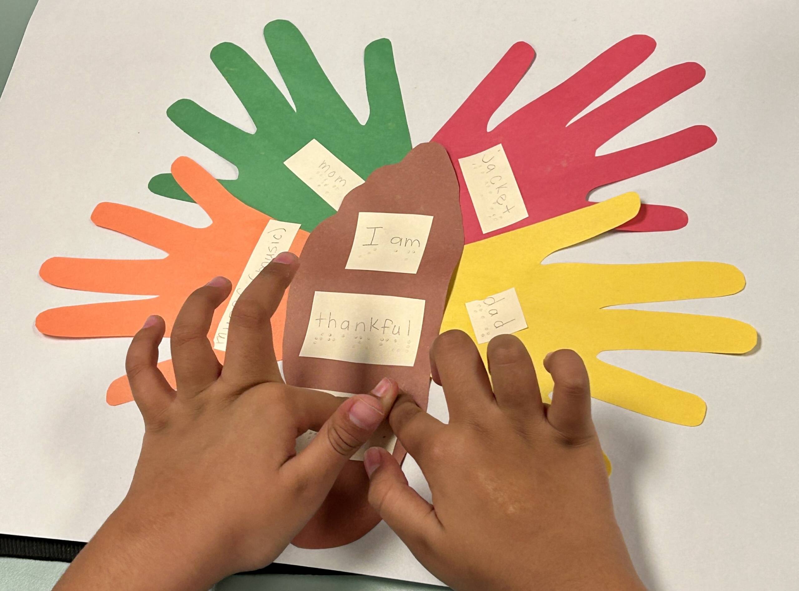 Thanksgiving placemat with cut out paper hands and a cut out foot in the middle with a child's hands touching the braille words on them. It is What They are Thankful for...Mom, Dad, dog