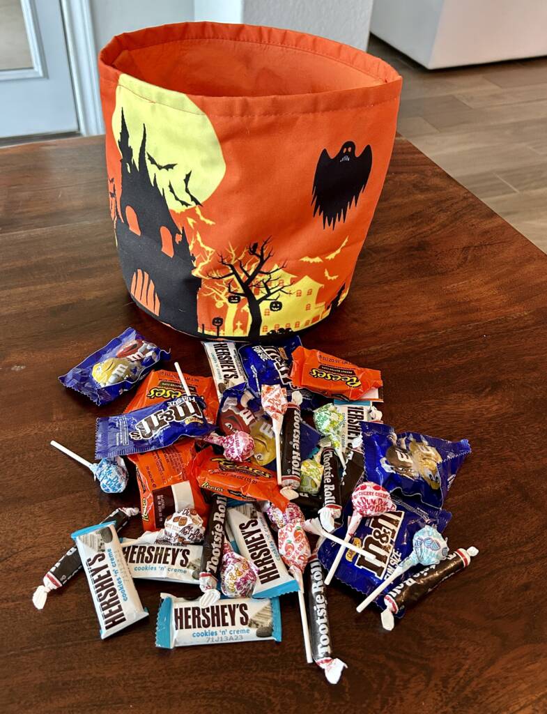 A pile of halloween candy on a table with a halloween bag behind it.
