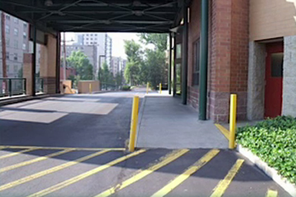 Diagonal yellow lines are painted on a section of the road. Two of those lines run up to vertical yellow posts that designate the sidewalk. In addition, dark shadows alternate with areas of bright sunlight.