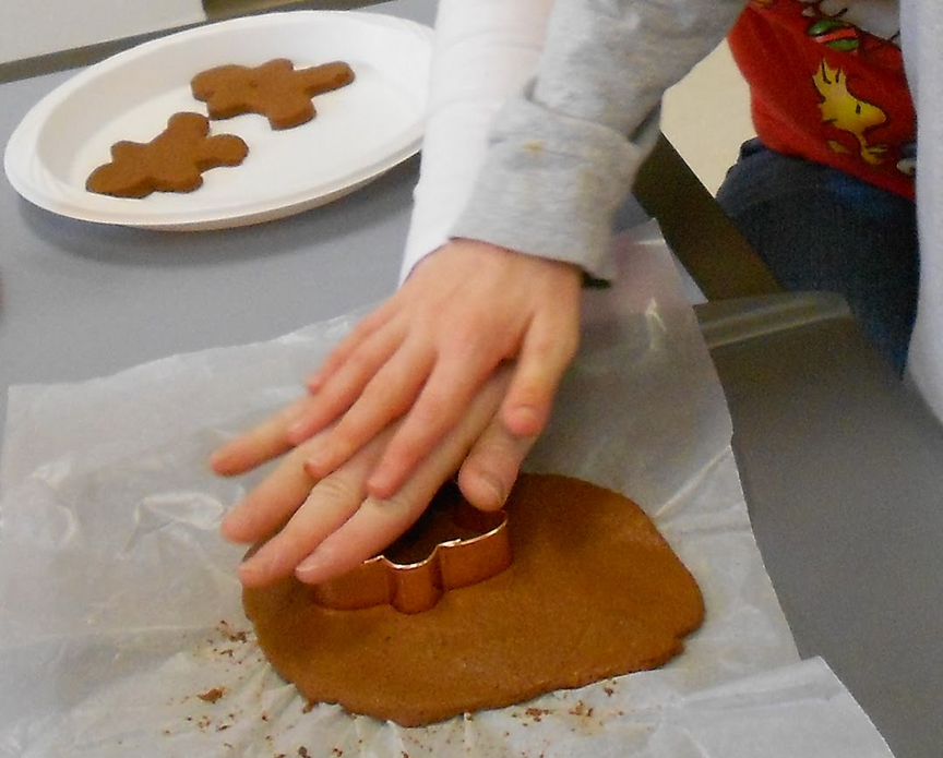 A teacher's hand with a child's hand on top of it pressing a gingerbread man cookie cutter into the dough
