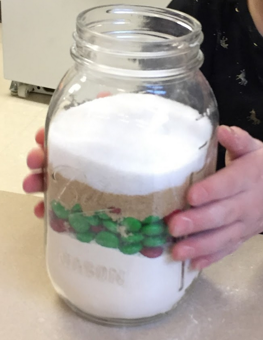 A mason jar with layers of ingredients to make cookies such as sugar, flour, and colored chocolate candies