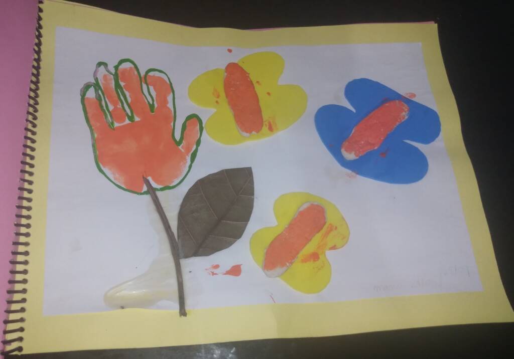 colored image of a sheet of paper containing the painting of the child left hand and underneath a dry branch with a dry leaf representing a flower made in the shape of a hand. Next to it are three colorful butterflies made with modeling clay.