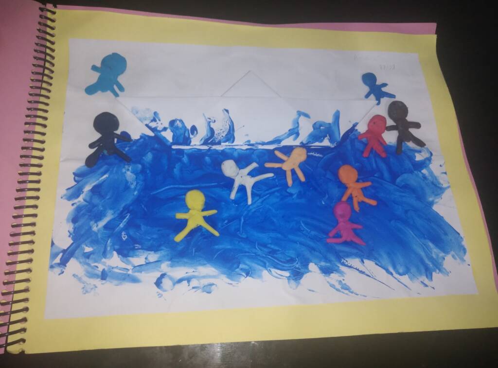 colored image of a sheet of paper containing a boat made of paper and 10 small dolls made with modeling clay of different colors. The paper boat and dolls are glued on top of a painting made with blue paint to represent the river.