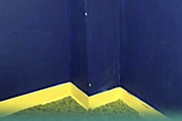 In a photograph, we see dark blue walls that come together in the corner of a classroom. A wide strip of bright yellow plastic that runs along the bottom of the walls clearly distinguishes the walls from the dark green carpeting on the floor.