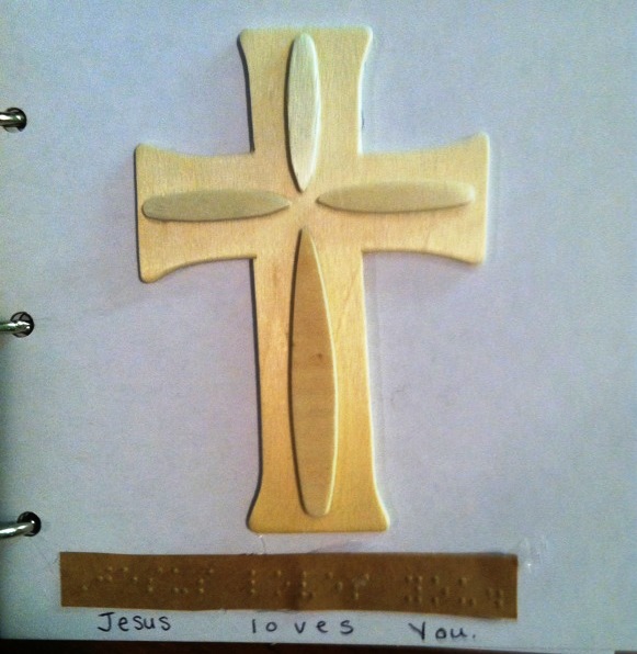 Wooden cross with text in print and braille, "Jesus loves you."