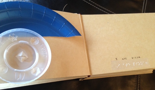Pages with plastic cup lid and part of plastic plate with braille text "I ate pizza."