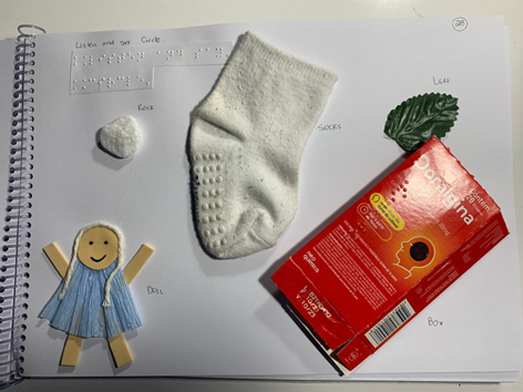 A page of a book with a paper doll, rock, sock, leaf, and box taped to the paper