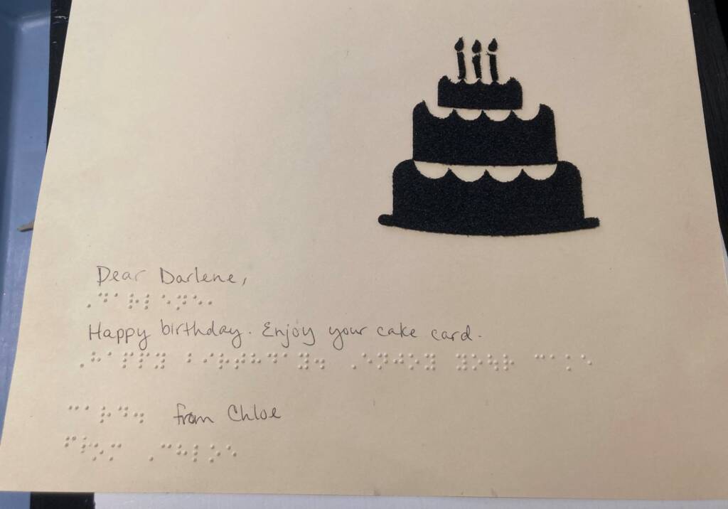 A greeting card with a cake on it and the words "Dear Darlene, Happy birthday. Enjoy your cake card. From Chloe" written in both print and braille.
