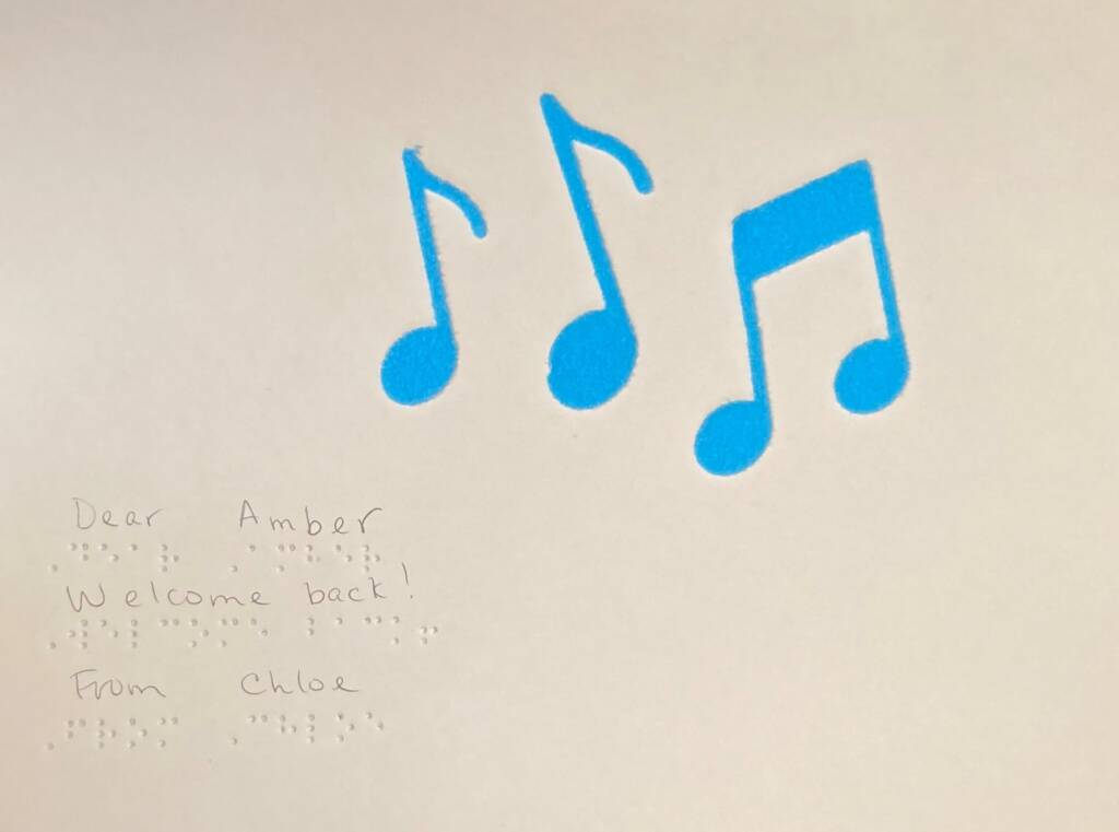 A piece of paper with three music notes on it and the words "Dear Amber, Welcome back! From Chloe" written in print and in braille.