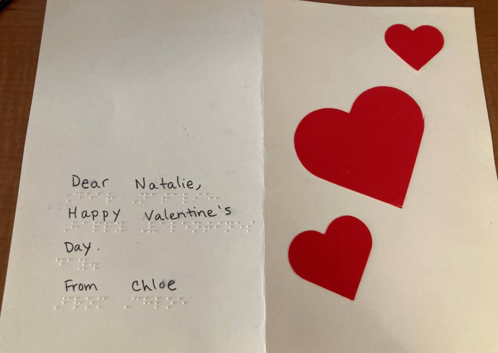 A greeting card that has three hearts on it and says "Dear Natalie, Happy Valentine's Day. From Chloe" in both print and braille.