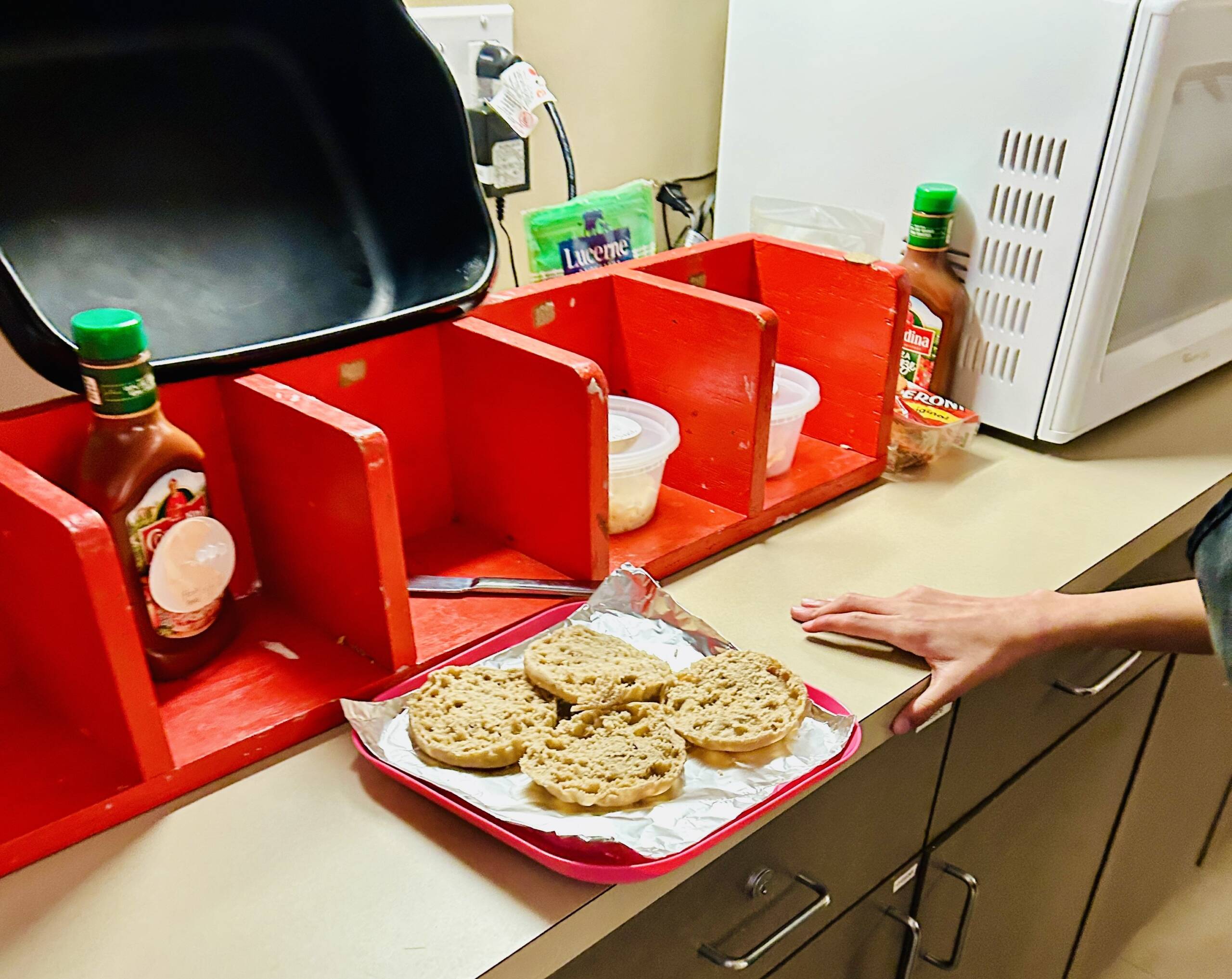 A countertop with a sequence box of the pizza ingredients in them (sauce, knife, cheese, toppings), and two English muffins in front of it