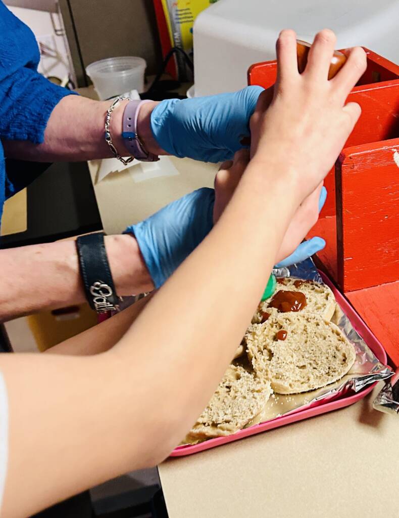 A student squeezing the pizza sauce onto the English muffins with the assistance of a teacher
