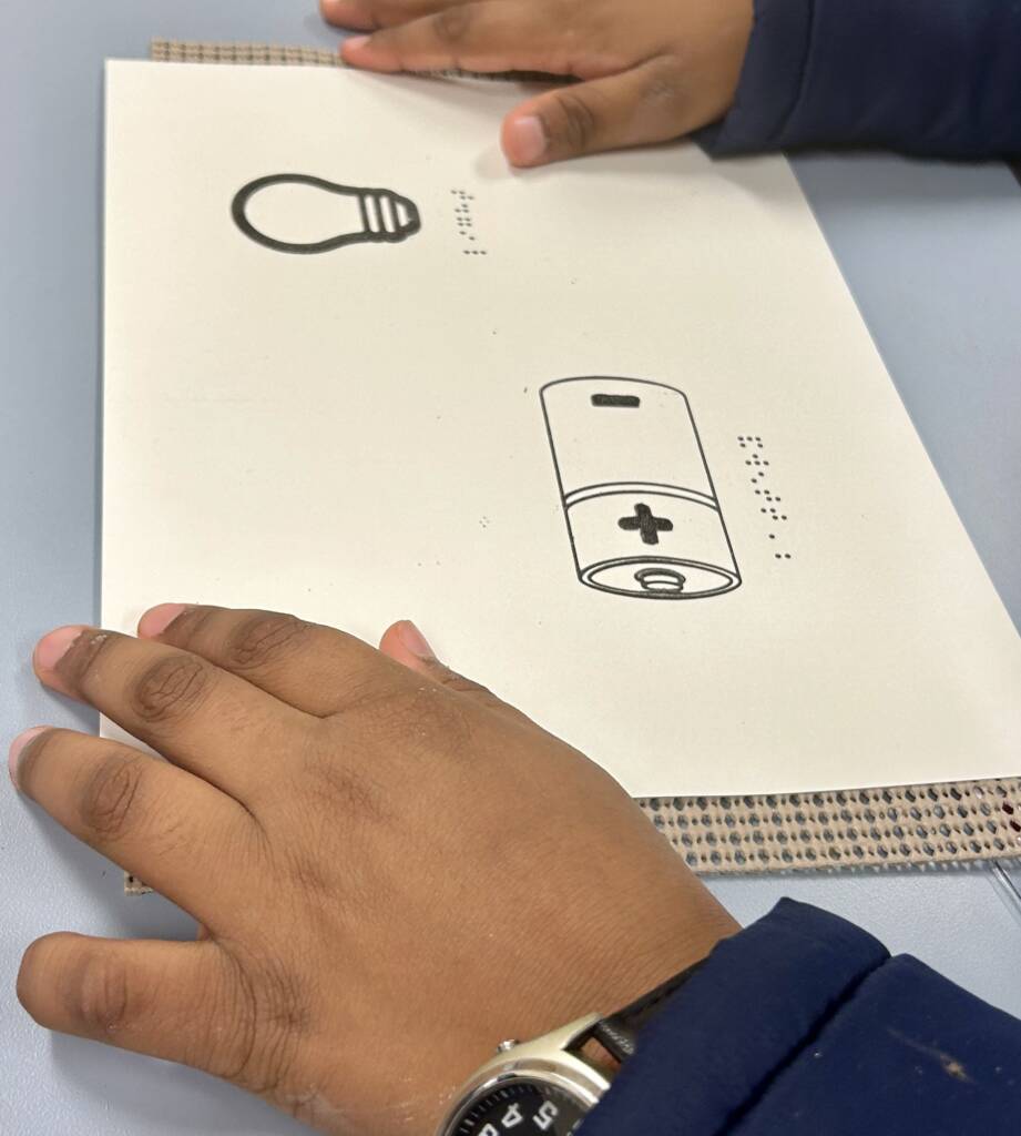 The circuit activity tactile graphic, which is a piece of paper with a drawing of a light bulb and battery on it, with braille written underneath each image.