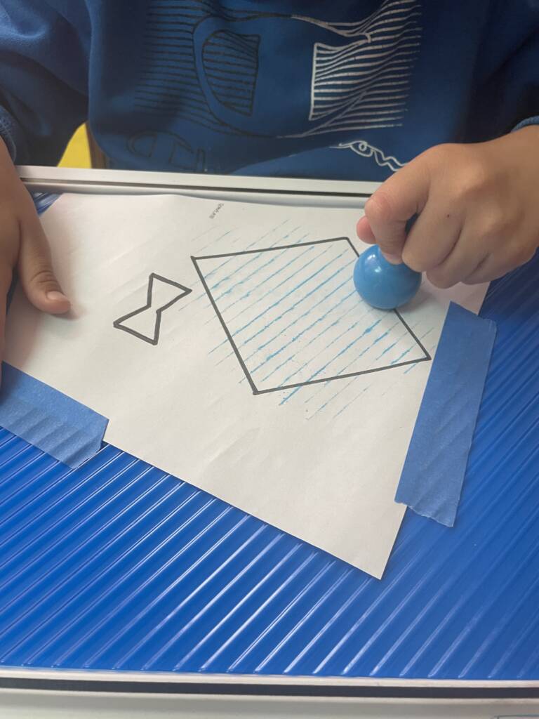 Student coloring the kite that is a raised line drawing over a bumpy mat and using an adapted crayon.