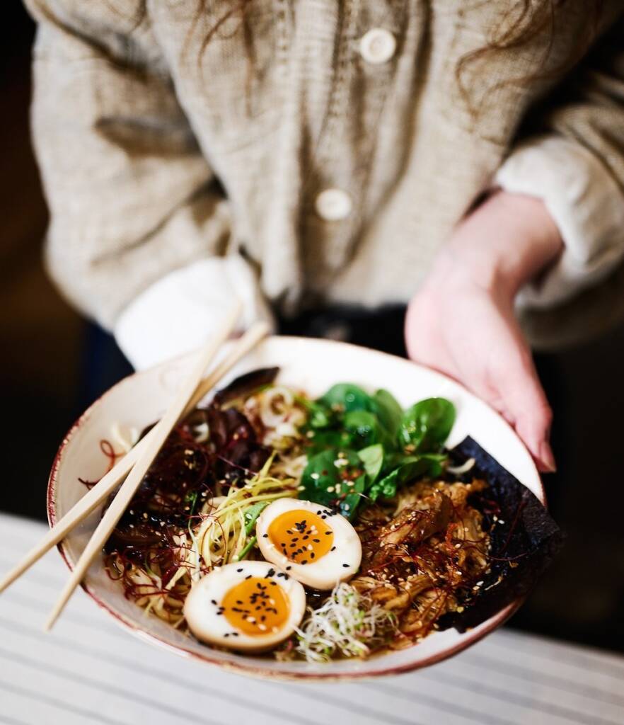Woman holding an asian dish that has ramen, an egg, and veggies.