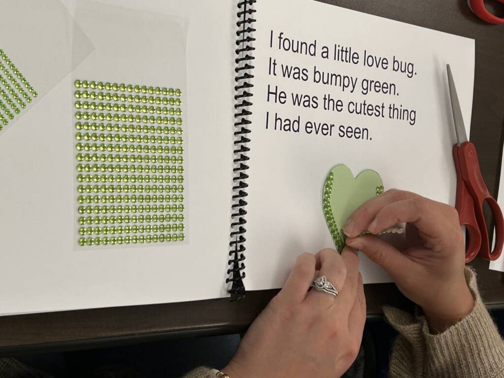Teacher making a page of the book with a green heart and craft gems for texture.