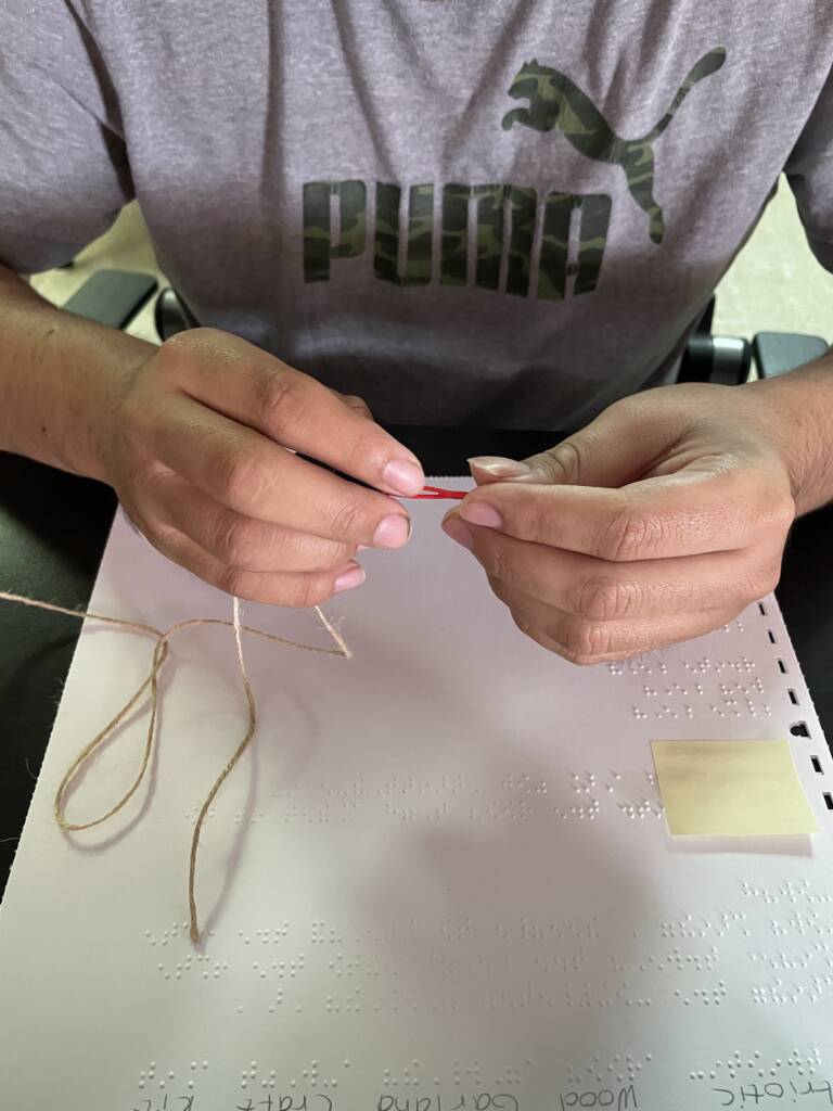 Student stringing bead onto string with braille directions on their table top.