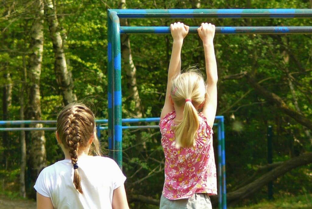 Two girls playing at the playground, one is hanging by her hands from a metal bar on the equipment.