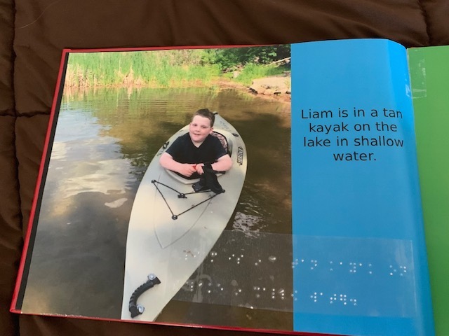 A photo of a page in a summer journal made by a student. The picture shows the boy sitting in a kayak and displays the following text written in both print and braille: "Liam is in a tan kayak on the lake in shallow water."