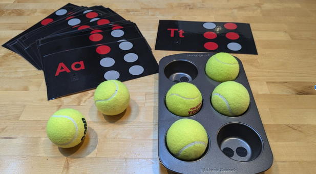 A stack of ABC braille cards with the muffin tin and tennis balls to make the braille cells.