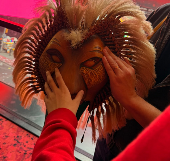 A child reaches out to touch an intricate Lion King mask, which is being held by an adult. The mask features a carved lion face with detailed textures and a mane made of woven and feathered materials. The background shows a large window overlooking Times Square, with bright red billboards and city buildings visible.