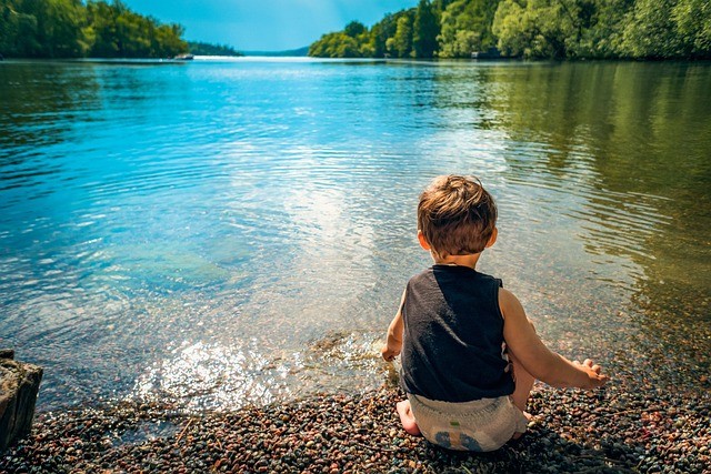 Young boy at the edge of a lake in bare feet touching the water.