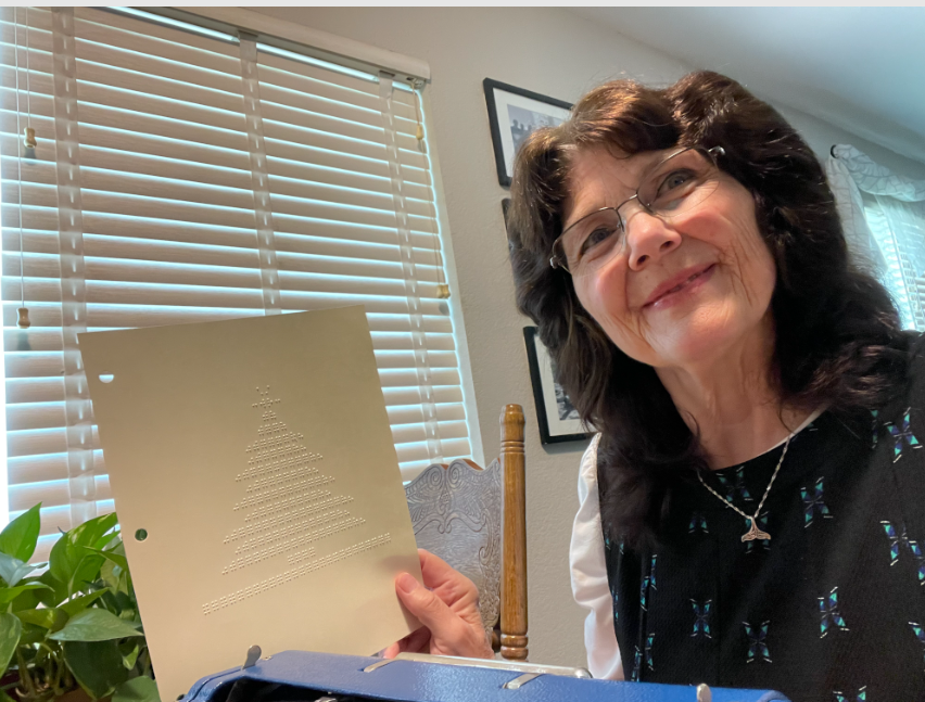 Edith at her braille machine smiling and holding a braille art design of the Christmas tree.