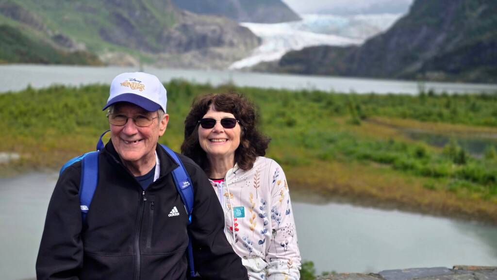 Edith and her husband in front of an Alaskan mountain that has a partially frozen river in the background.