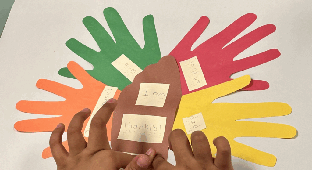 Placemat with paper hands and braille with print that says: I am thankful for mom, dad, Jackey with student hands touching the braille.