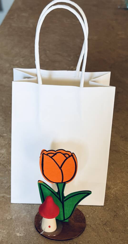 A completed orange painted wooden flower in front of a small, handle paper bag. 