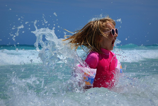 Girl jumping in a wave with sunglasses, floaties, and a big smile.