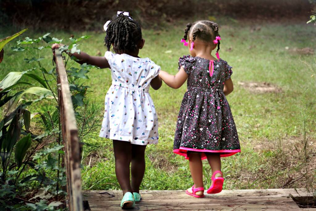 Two little girls in pretty summer dresses holding hands and crossing a little wooden bridge. 