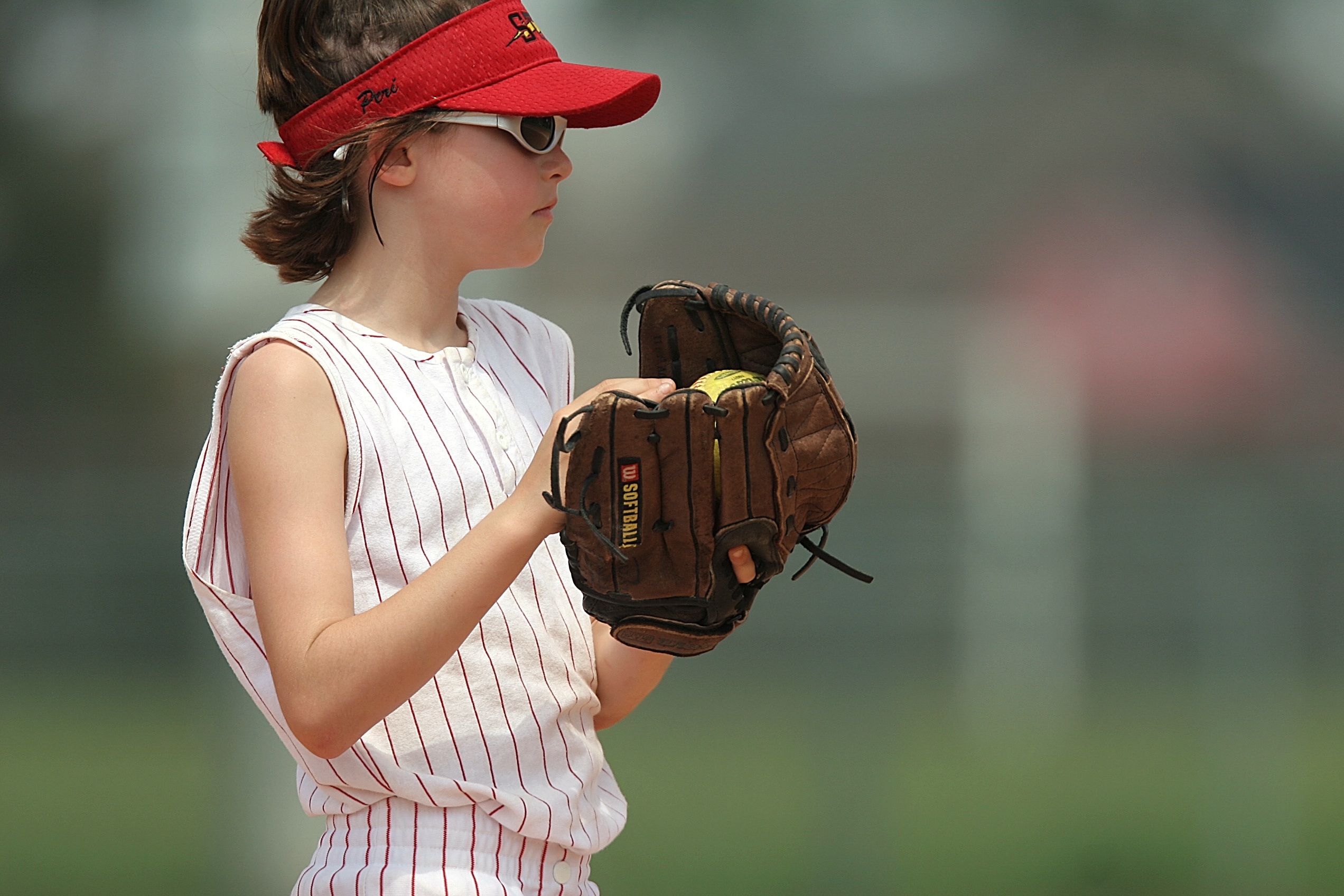 Young girl with a sunvisor on and sunglasses holding a baseball and mit.