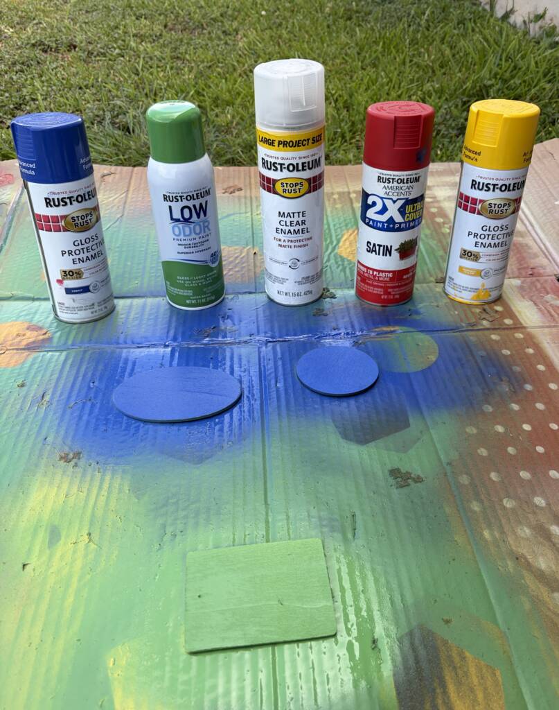 Table with spray paint cans and wooden circle and square pieces that are being sprayed since colors that include blue and green.