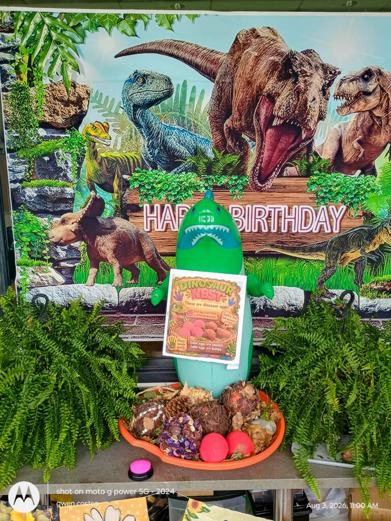 Fossil nest station with a large poster of dinos as the background, ferns on each side, a blowup dino with an information sheet, and a talking AAC switch in front of a constructed dino nest exhibit.