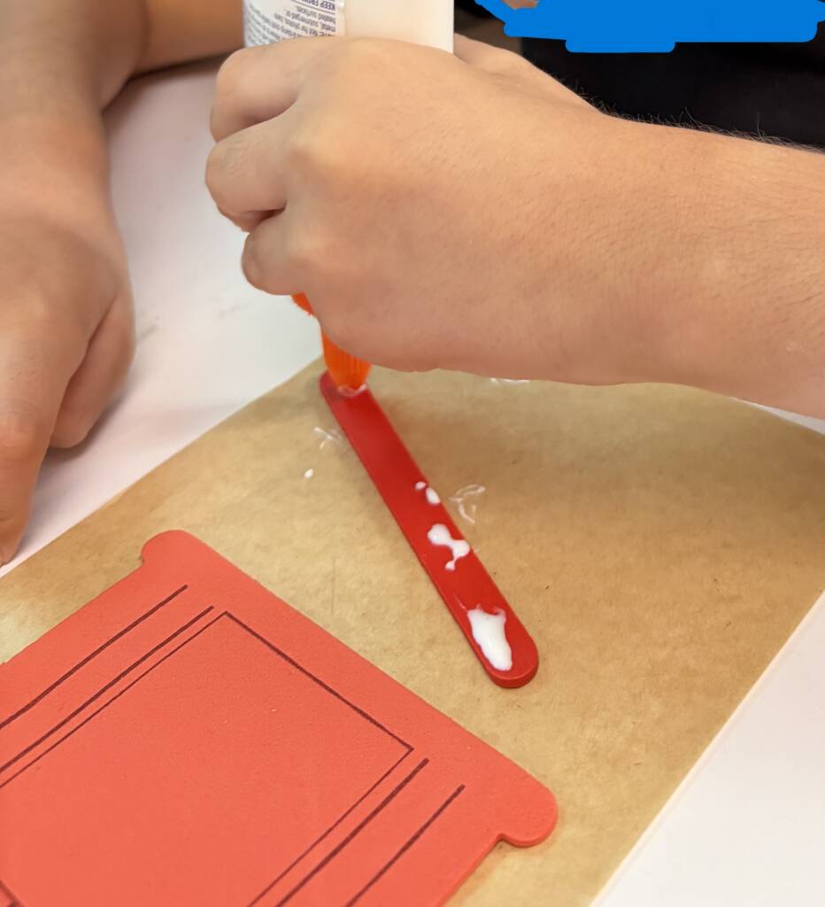 Student glueing a red popsicle stick as part of their barn craft.