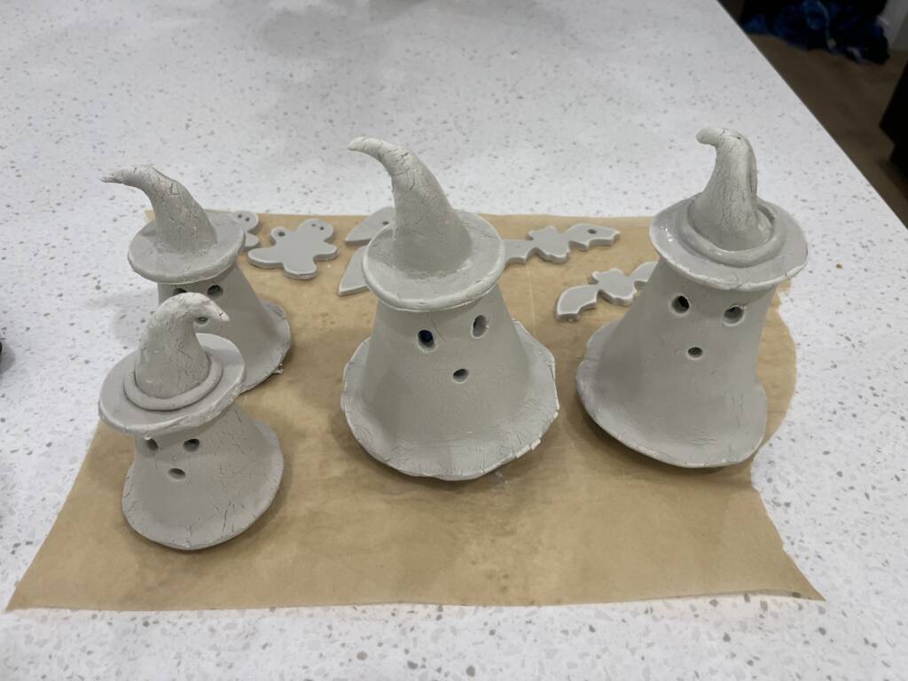 Two large and two smaller clay ghosts with witch hats drying and a few cookie cutter clay bat ornaments behind them. 