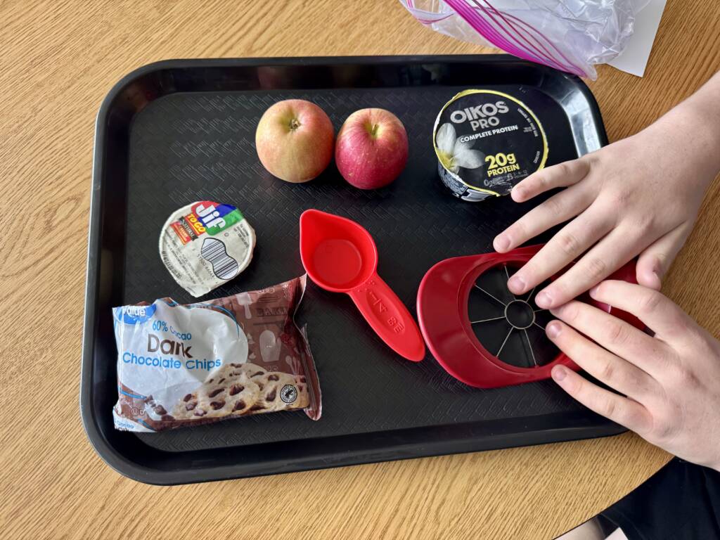 Tray with Liam touching the following items: Measuring cups (braille of course), bowl, vanilla Greek yogurt, dark chocolate chips,
peanut butter, spoon, apples, apple cutter.