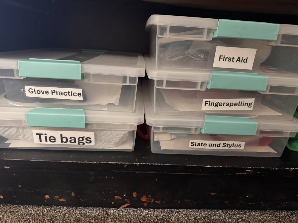 Labeled boxes that include glove practice, tie bags, first aid, fingerspelling, slate and stylus.