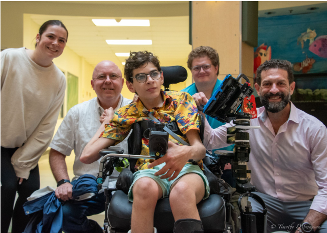 LJ with some of his team members around him smiling. LJ has his accessibility features mounted on his chair for ease of access.