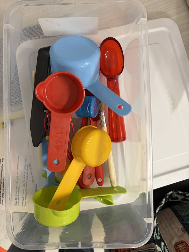 Kitchen bin with various small tool kitchen items that include measuring cups, whisk, and spoon.